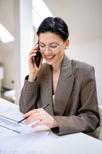 Femme d'affaires confiante portant des lunettes, parlant au téléphone tout en examinant des documents dans un environnement de bureau moderne.