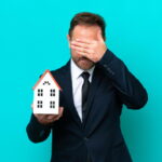Un homme en costume, l’air inquiet, tenant une petite maquette de maison d'une main et se couvrant le visage de l'autre, illustrant le désarroi lié aux vices cachés et litiges après un achat immobilier.