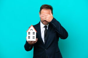 Un homme en costume, l’air inquiet, tenant une petite maquette de maison d'une main et se couvrant le visage de l'autre, illustrant le désarroi lié aux vices cachés et litiges après un achat immobilier.