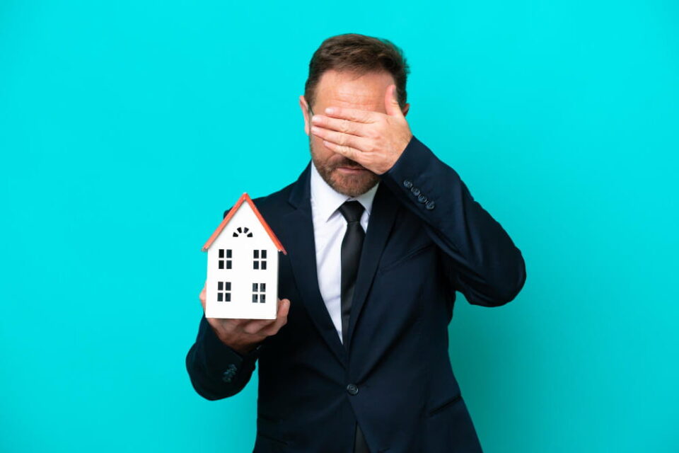 Un homme en costume, l’air inquiet, tenant une petite maquette de maison d'une main et se couvrant le visage de l'autre, illustrant le désarroi lié aux vices cachés et litiges après un achat immobilier.